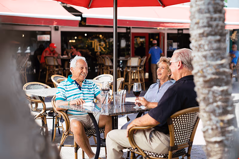Three older adults sit at a patio table under a red umbrella at an outdoor café, chatting and holding drinks.