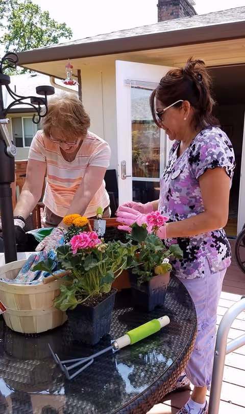 Two women gardening on a patio table with potted flowers and gardening tools. One woman is wearing a striped shirt and glasses, and the other is wearing a purple patterned shirt and pink gloves. They are working together with soil and plants in a sunny outdoor setting.