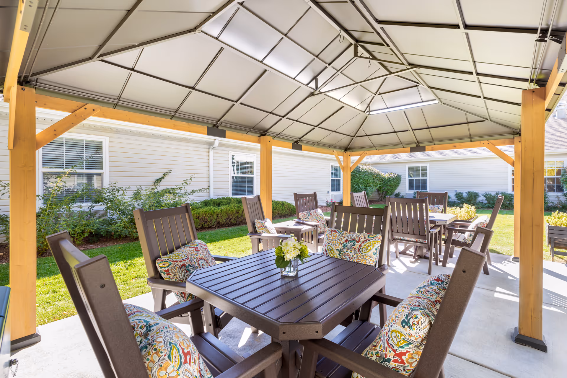 Outdoor covered patio area with multiple dark brown tables and chairs, each chair featuring colorful patterned cushions. The patio has a metal roof supported by wooden beams, and is surrounded by green grass and bushes with a beige building in the background.