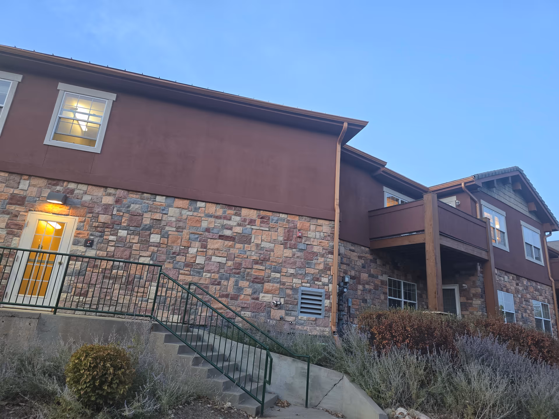 Exterior view of a two-story residential building with a stone lower facade, balcony, exterior stairs and a lit doorway under a clear sky.