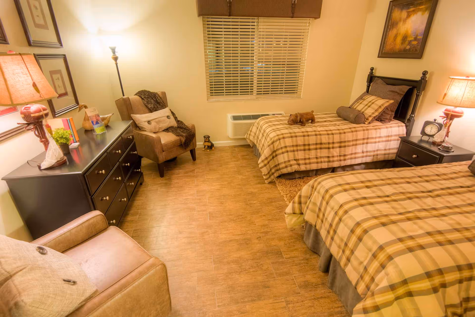 A cozy bedroom with two twin beds covered in plaid bedding. There is a window with blinds on the far wall, a dark wooden dresser with a lamp and decorative items, and two armchairs with cushions. The room has warm lighting and wooden flooring.