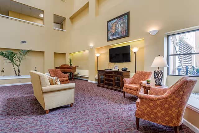 A spacious living room area in a senior living facility with beige walls and high ceilings. The room features a beige sofa, two patterned armchairs, a wooden TV stand with a flat-screen TV mounted above it, two floor lamps, a table lamp on a side table, a piano in the corner, and a large window letting in natural light. There is a framed painting hanging above the TV.