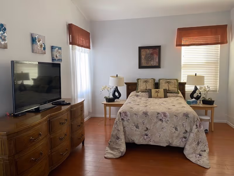 A bedroom with a bed covered in a floral bedspread and three decorative pillows. There are two wooden nightstands on either side of the bed, each with a lamp and a small plant. A wooden dresser with a flat-screen TV on top is positioned to the left. The room has two windows with brown valances and white blinds, and light-colored walls with a framed artwork above the bed.