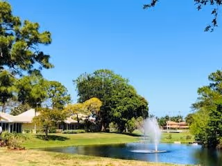 A scenic outdoor view featuring a pond with a water fountain in the center, surrounded by green grass, trees, and residential buildings under a clear blue sky.