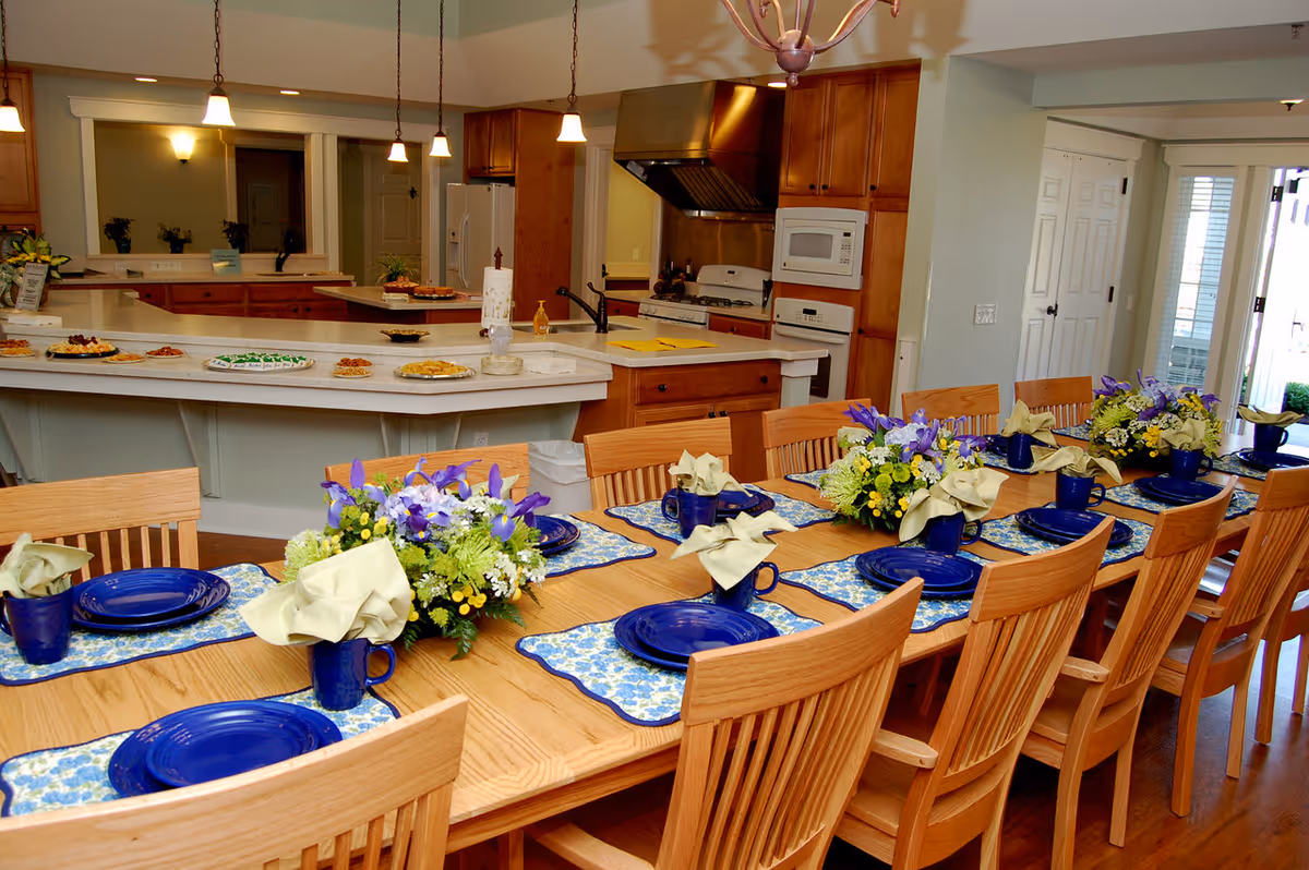 A long wooden dining table set with blue plates, matching blue mugs with beige napkins, floral placemats, and flower centerpieces. The table is surrounded by wooden chairs. In the background, there is a kitchen area with countertops, cabinets, a stove, microwave, and refrigerator. Various dishes are placed on the kitchen counter.