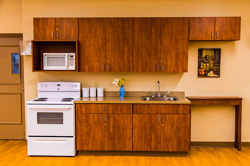 Interior view of a kitchen area with wooden cabinets, a white electric stove, a microwave, a countertop with a sink, a blue vase with flowers, and a small wooden table against a beige wall.