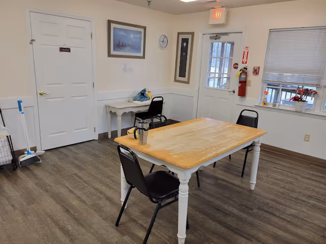 A small room with a wooden table and three black chairs around it. There is a stainless steel travel mug on the table. The room has light-colored walls with a white door labeled 'Cleaning', a window with blinds, a fire extinguisher mounted on the wall, and a small white desk with a chair. The floor is covered with wood-patterned vinyl or laminate flooring. There are framed pictures and a clock on the walls.