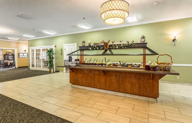 Interior view of a senior living facility dining area with a buffet counter featuring various food items and utensils. The room has light green walls, a hanging light fixture, and a carpeted area leading to a room with glass double doors.