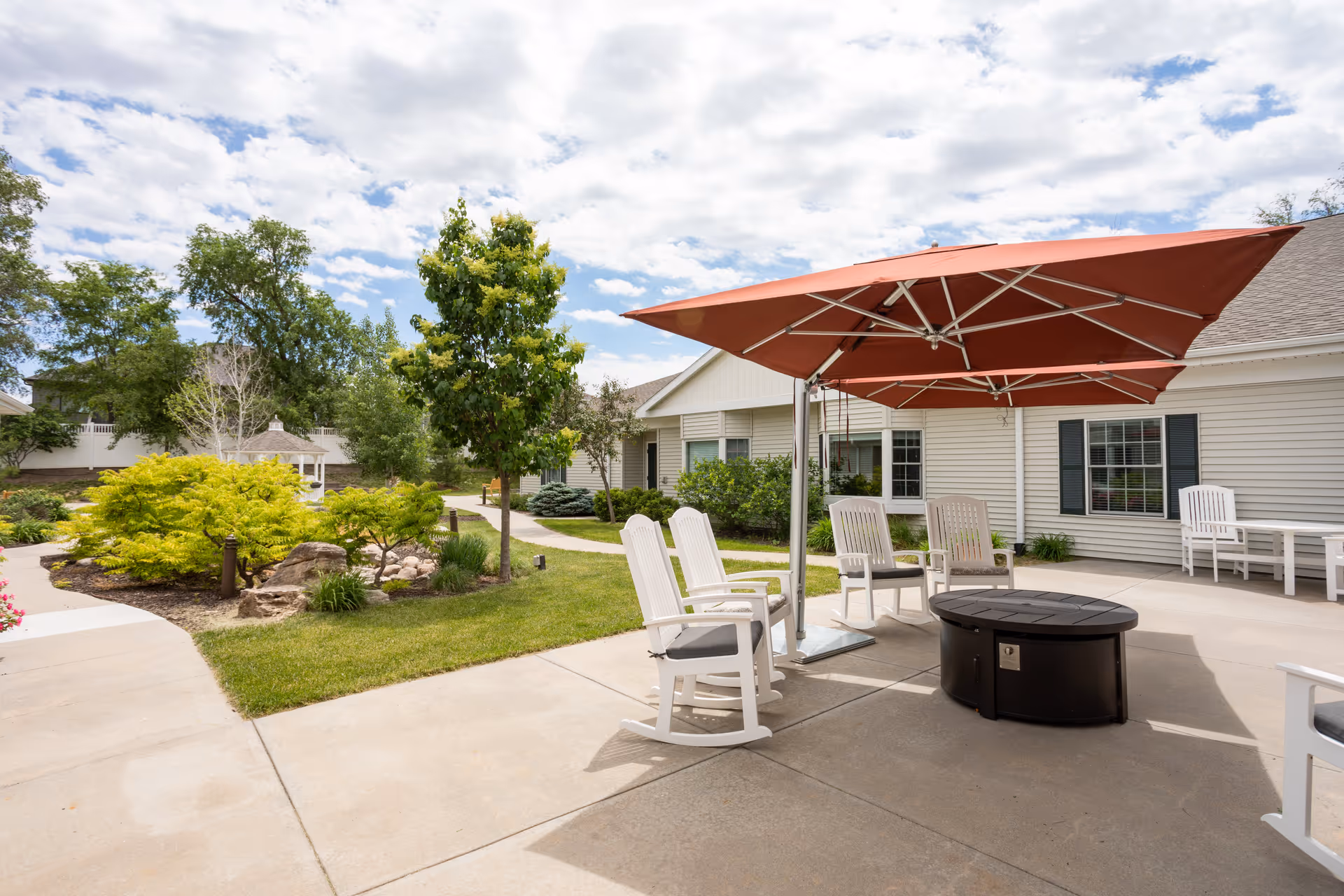 Outdoor patio area at CountryHouse Residence featuring white rocking chairs arranged around a circular fire pit table under a large red umbrella. The patio is surrounded by a well-maintained garden with green shrubs, trees, and a walkway leading to a white building with windows and black shutters. The sky is partly cloudy.
