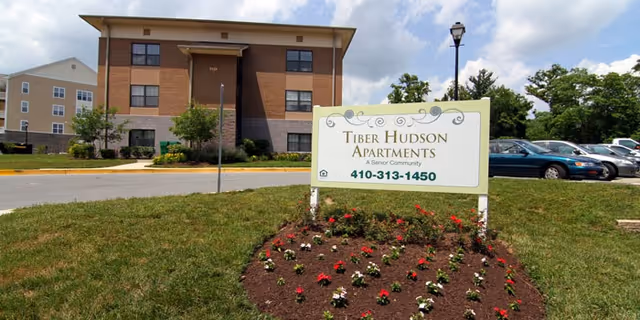 A landscaped sign reading "Tiber Hudson Apartments" stands in front of a three-story apartment building and parked cars.