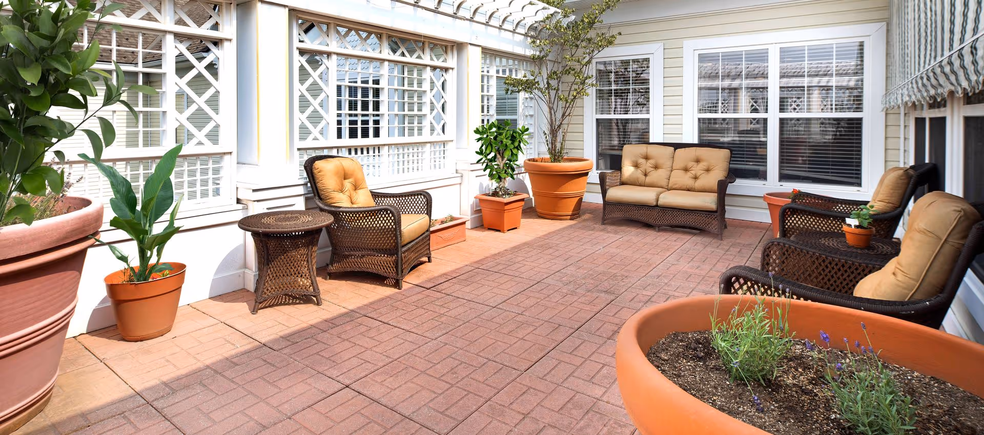 Sunlit outdoor patio with wicker chairs, a cushioned loveseat, side tables and large potted plants along a white lattice wall.