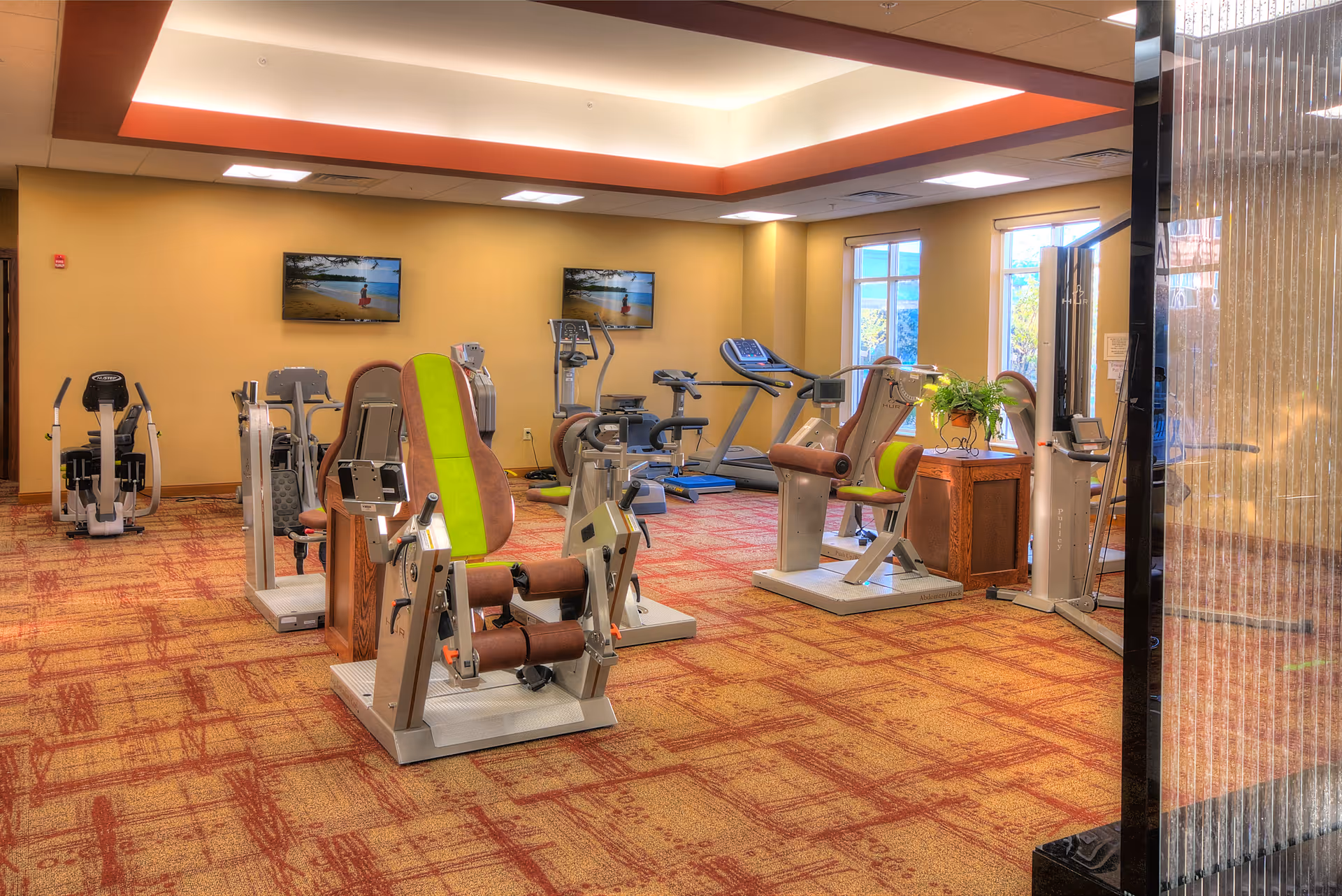 Well-lit fitness room with seated resistance machines, treadmills, and wall-mounted TVs on a patterned carpet.