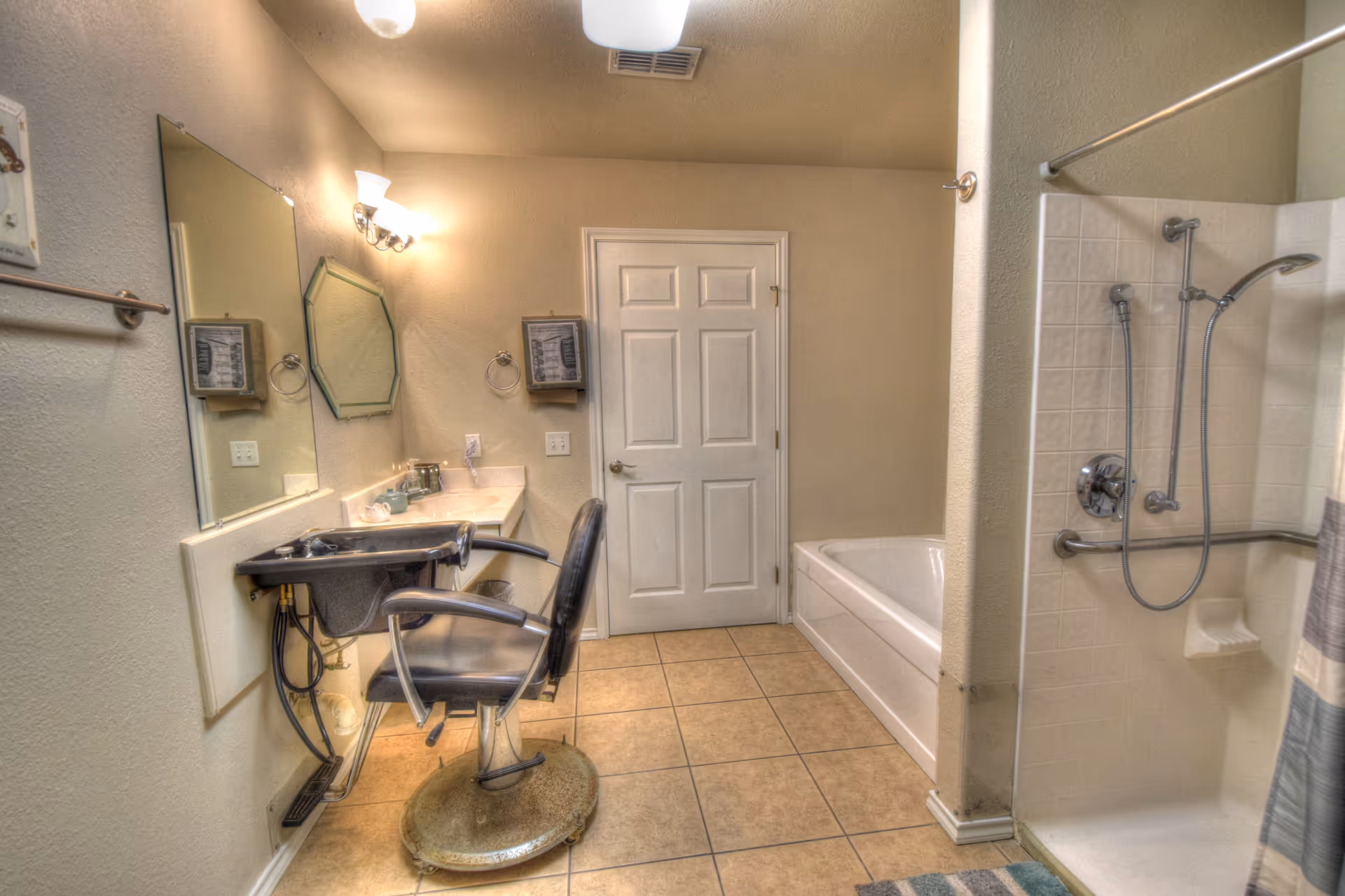 A bathroom with beige tiled floor and walls painted in light beige. On the left side, there is a large mirror above a counter with a sink and a black salon-style chair in front of it. The back wall has a white door, and to the right, there is a white bathtub. On the far right, there is a walk-in shower with a handheld showerhead and grab bars. The room is well-lit with wall-mounted lights above the mirror.