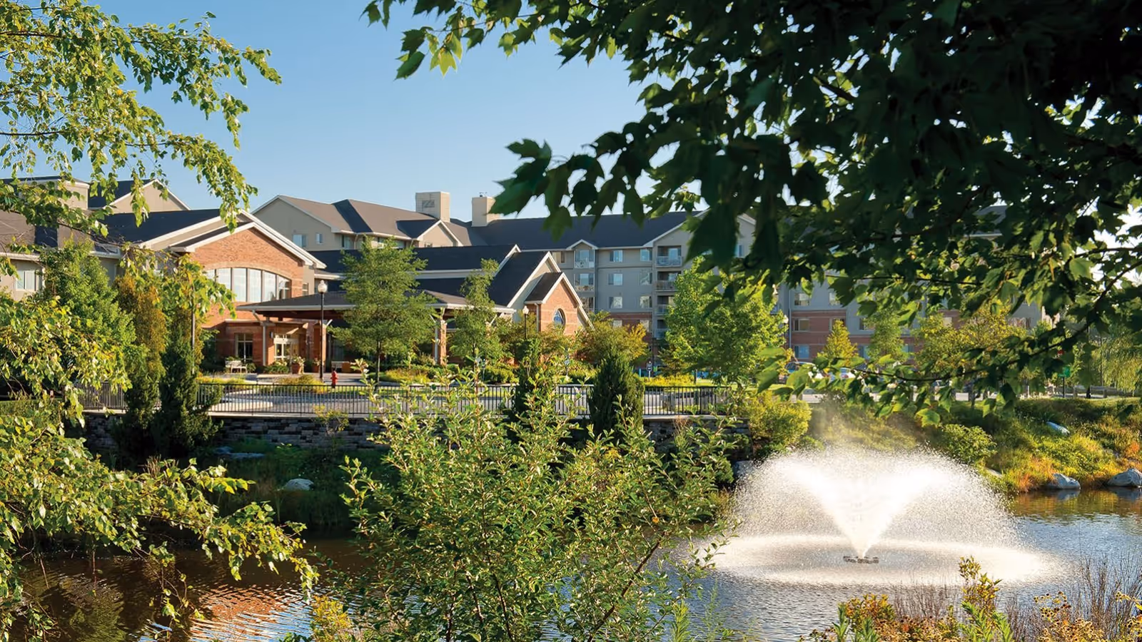 A landscaped pond with a central fountain in front of a multi-story senior living building and patio, framed by leafy trees.