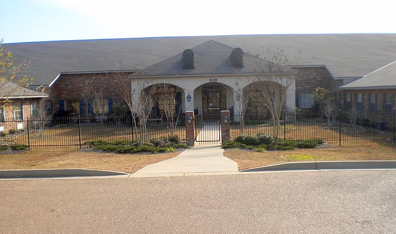 Front exterior view of a single-story brick building with a central entrance featuring an arched porch and a gated walkway leading to the door. The building has a sloped roof and is surrounded by a low fence and some landscaping with small trees and shrubs.