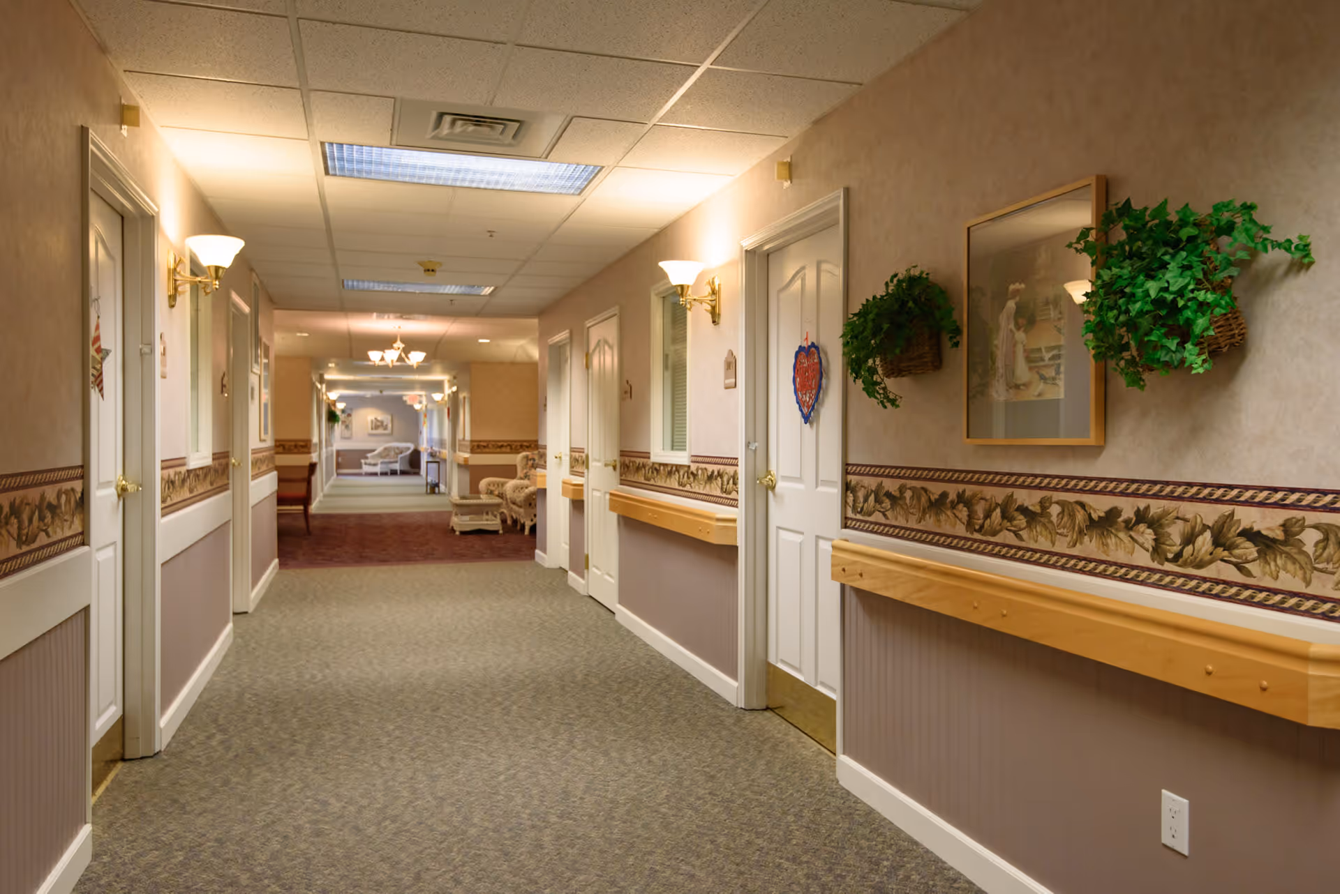 A well-lit hallway in a senior living facility with beige walls, decorative wallpaper border, and carpeted floor. Several white doors line both sides of the hallway, some adorned with small decorations. Wall-mounted light fixtures and hanging plants add to the decor. At the end of the hallway, there is a seating area with chairs and a table.