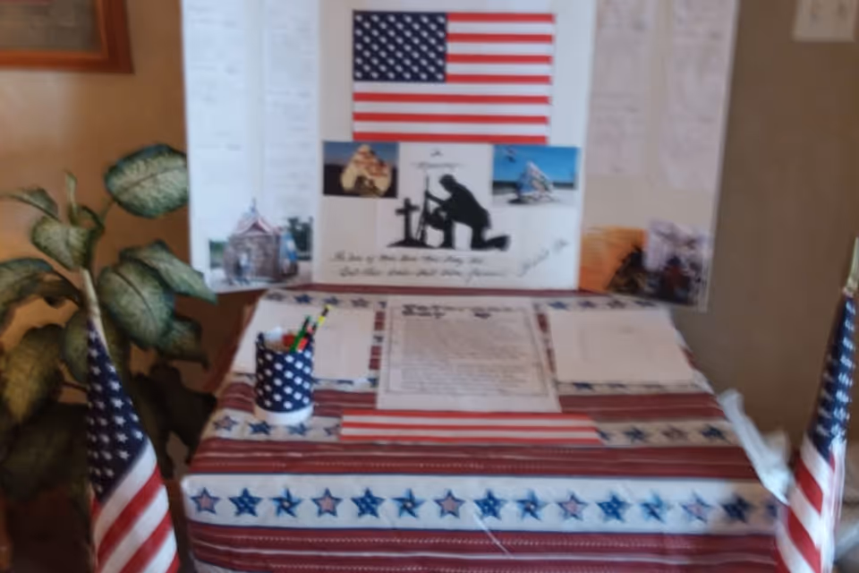 A patriotic display table covered with a red, white, and blue tablecloth featuring stars and stripes. On the table are papers and a cup holding pens. Behind the table is a board with an American flag, photos, and a silhouette of a soldier kneeling by a cross. Two small American flags are placed on either side of the table, and a green leafy plant is visible to the left.