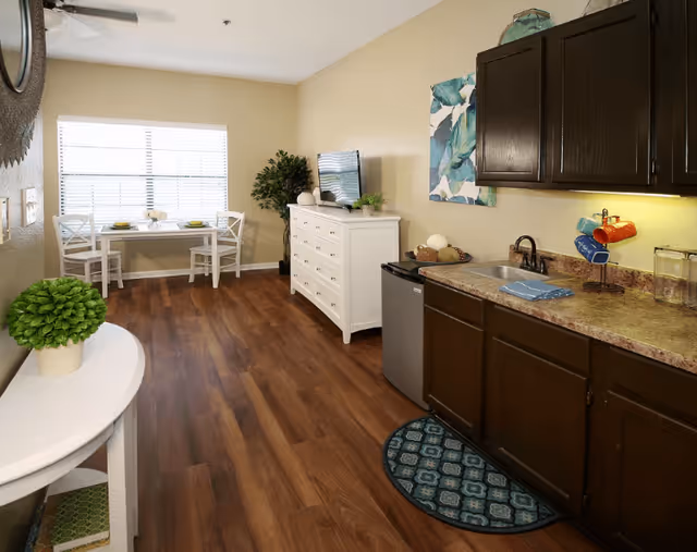 Interior view of a senior living facility room with a kitchenette on the right featuring dark wood cabinets, a small sink, a mini fridge, and a countertop with a few items. On the left side, there is a white console table with a green potted plant. In the background, a white dresser with a TV on top is against the wall, next to a large window with blinds and a small dining table with two chairs set with plates and cups. The room has wood flooring and light-colored walls with a decorative painting and a potted plant near the window.