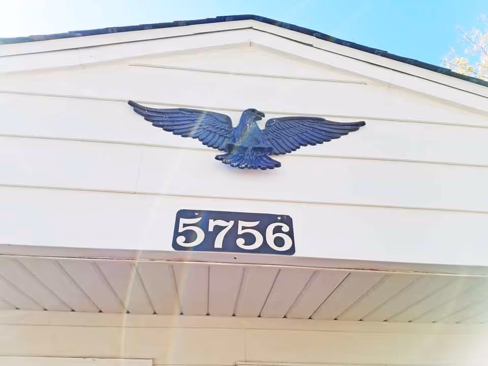 Close-up view of the front exterior of a building showing a white gable with a decorative blue eagle mounted above a black address plaque displaying the number 5756.