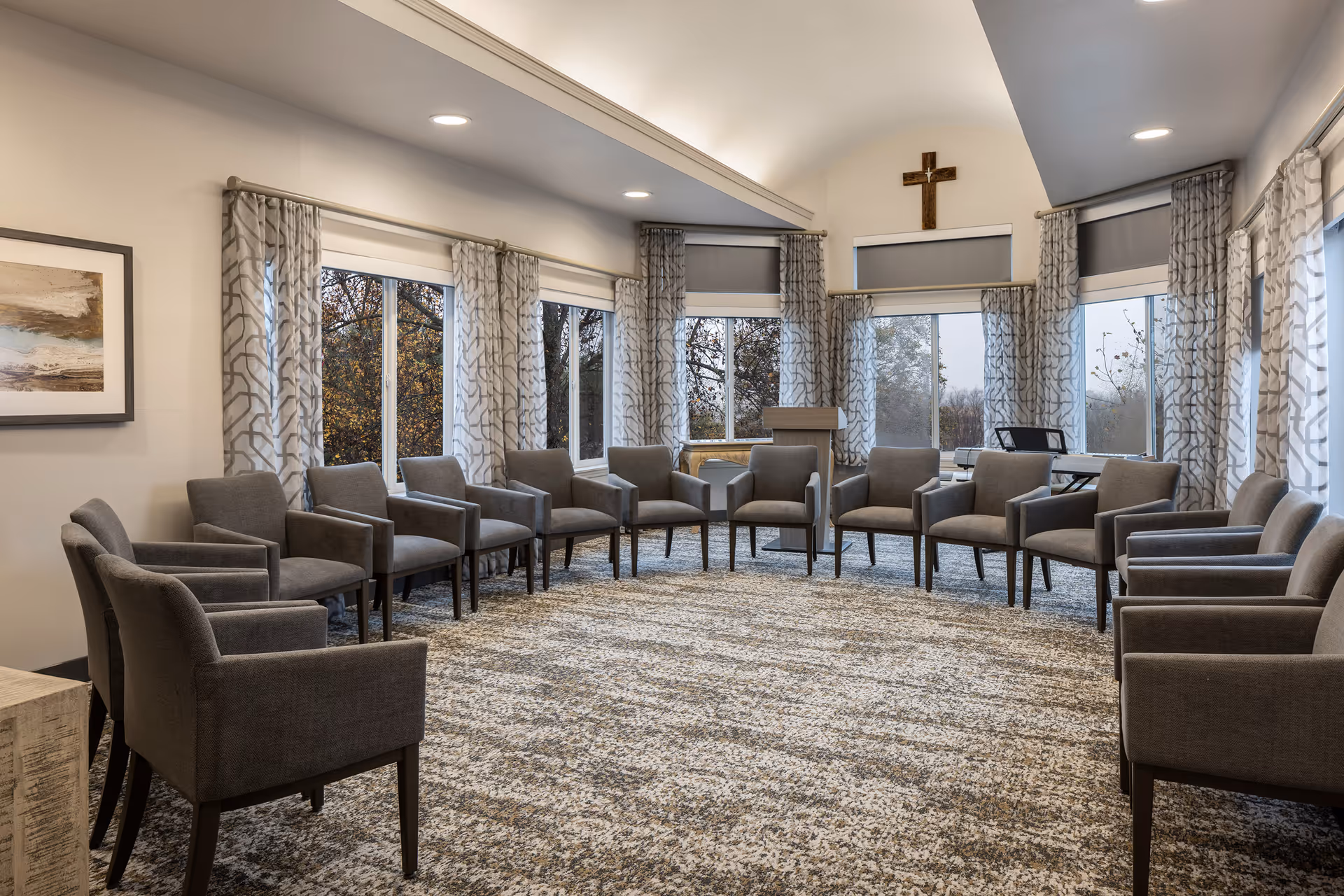 A spacious meeting or activity room with a circle of gray upholstered chairs arranged on a patterned carpet. Large windows with patterned curtains let in natural light, and a wooden podium stands near the windows. A cross is mounted on the wall above the windows.
