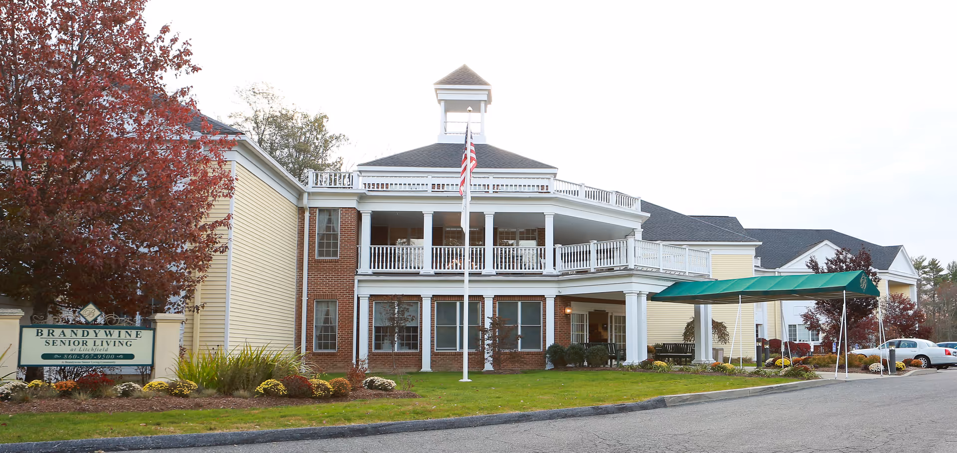 Two-story senior living building with white columns, brick accents, flagpole, and green entrance canopy.