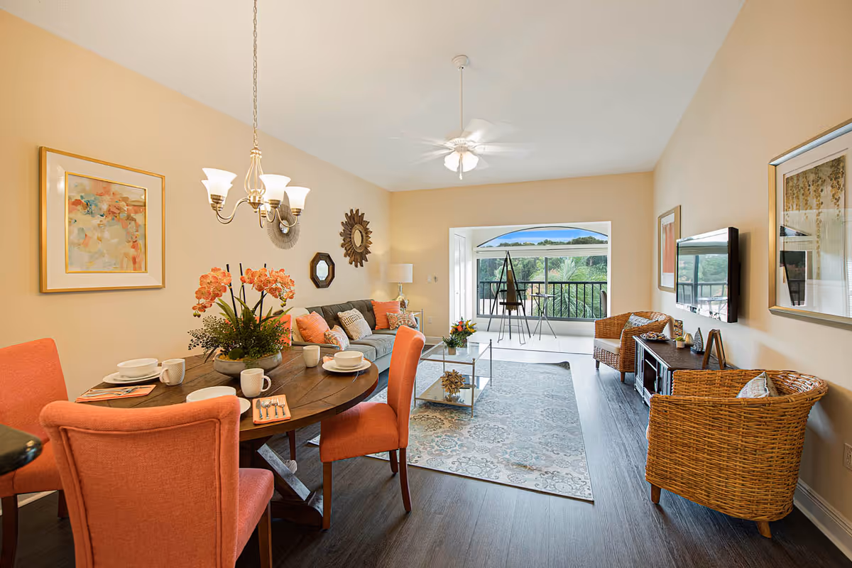 A bright open living and dining room with a round table and coral chairs, a sofa and wicker seating, coffee table, and sliding doors opening to a balcony overlooking greenery.