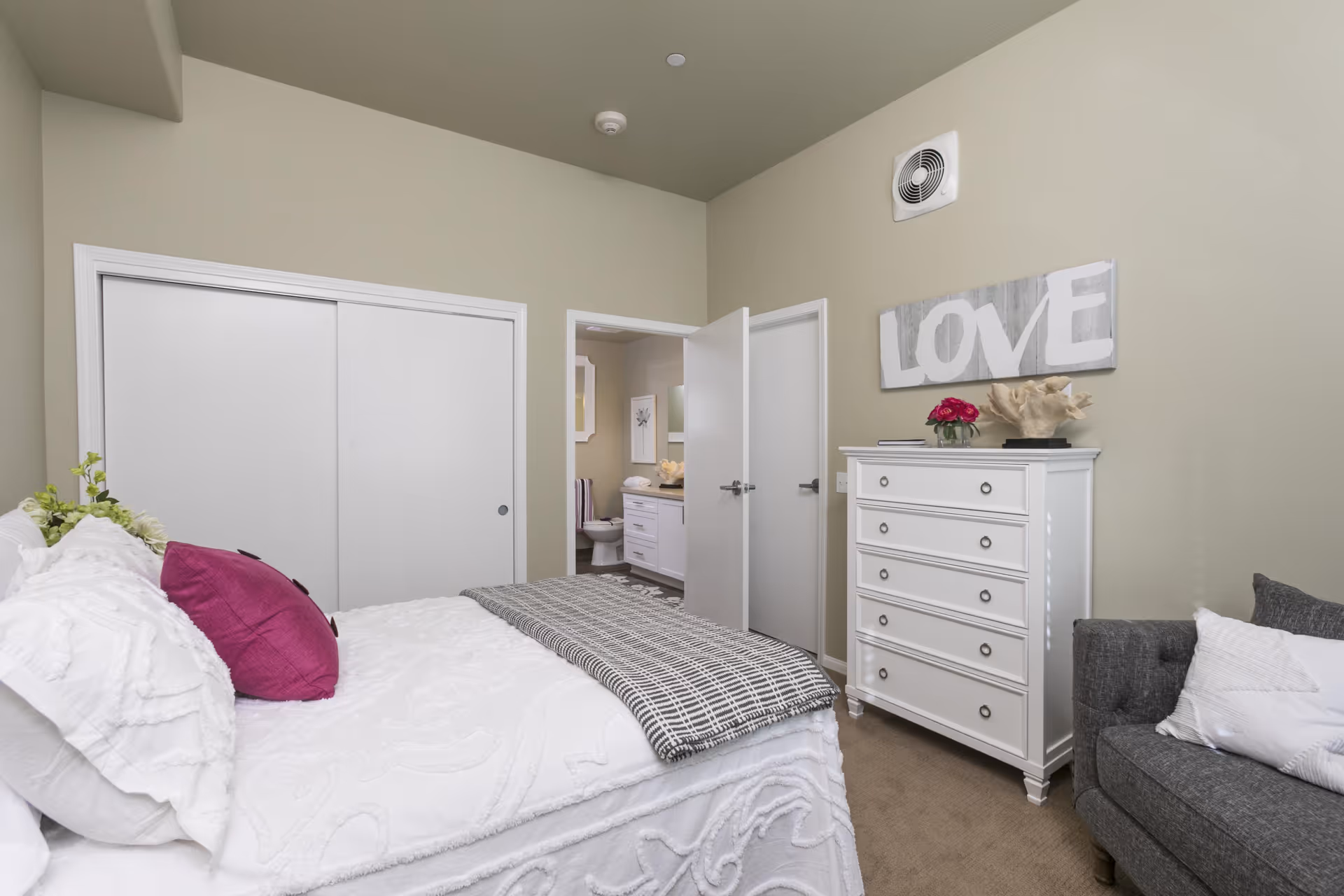 A bedroom with a white bedspread and a purple pillow, a white dresser with decorative items and a 'LOVE' sign above it, a gray armchair with a white pillow, and an open door leading to a bathroom with a toilet and vanity visible.