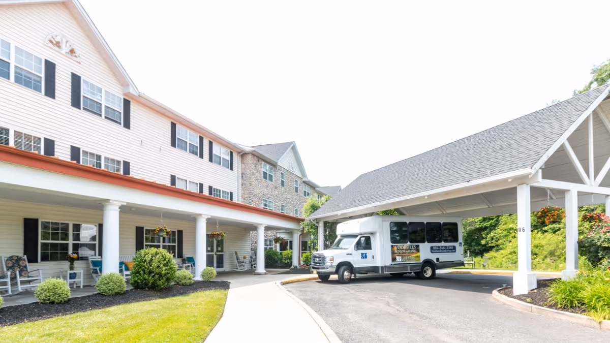 Exterior view of a senior living facility with a covered entrance and a shuttle bus parked under the canopy. The building has multiple windows, white columns, and a porch area with chairs and hanging plants. There is a well-maintained lawn and landscaping around the driveway.