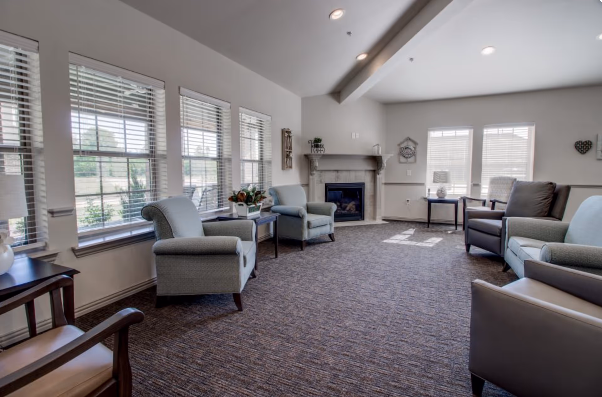 A bright and spacious living room with multiple armchairs arranged around a carpeted floor. Large windows with white blinds let in natural light. There is a fireplace with a mantel on the far wall, decorated with small plants and wall hangings. Side tables with lamps and decorative items are placed near the chairs.