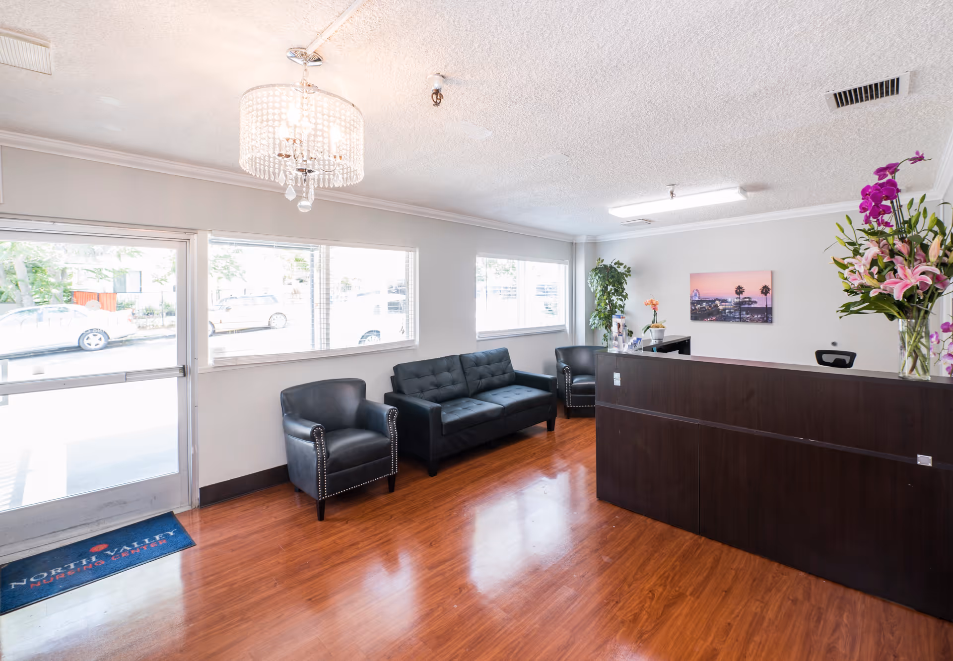 Reception area of North Valley Nursing Center with a dark wooden front desk, two black armchairs, a black sofa, large windows letting in natural light, a chandelier, and floral arrangements on the desk.