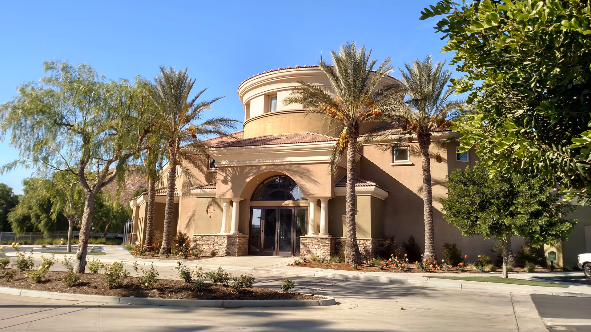 Exterior view of a senior living facility building with a beige stucco facade, arched entrance with columns, and a round upper section. The building is surrounded by palm trees, other greenery, and a clear blue sky.