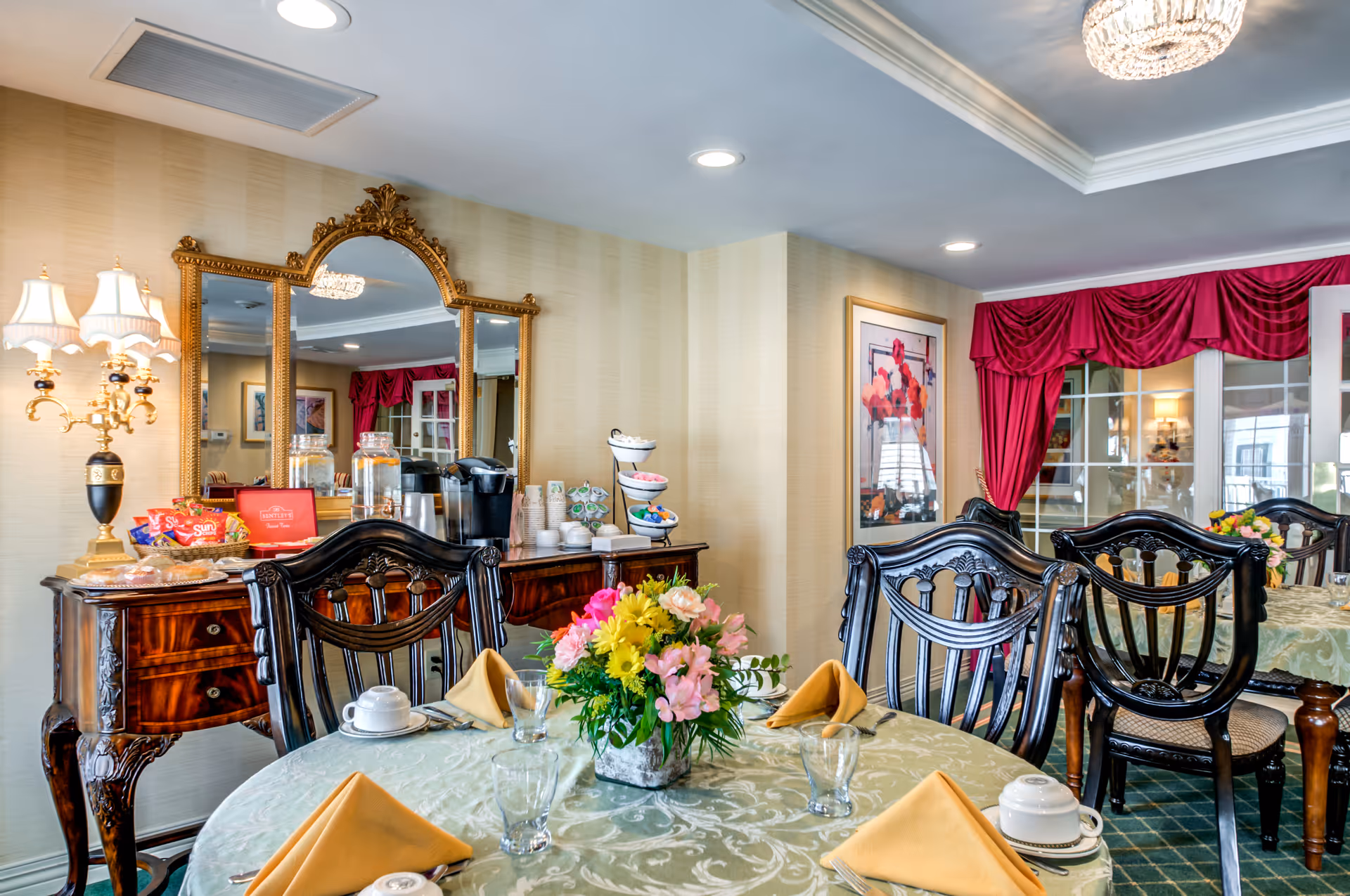 A dining room with a round table covered with a green patterned tablecloth, set with yellow folded napkins, cups, and glasses. A floral centerpiece with pink, yellow, and white flowers is in the middle of the table. Behind the table, there is a wooden sideboard with a large ornate gold-framed mirror above it, a coffee maker, snacks, and cups. The room has elegant dark wooden chairs, a chandelier on the ceiling, and red curtains on the windows.