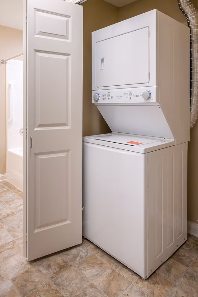 A stacked white washer and dryer unit in a small laundry closet with a partially open door. The floor has a tile pattern, and a bathroom with a bathtub and grab bar is visible in the background.