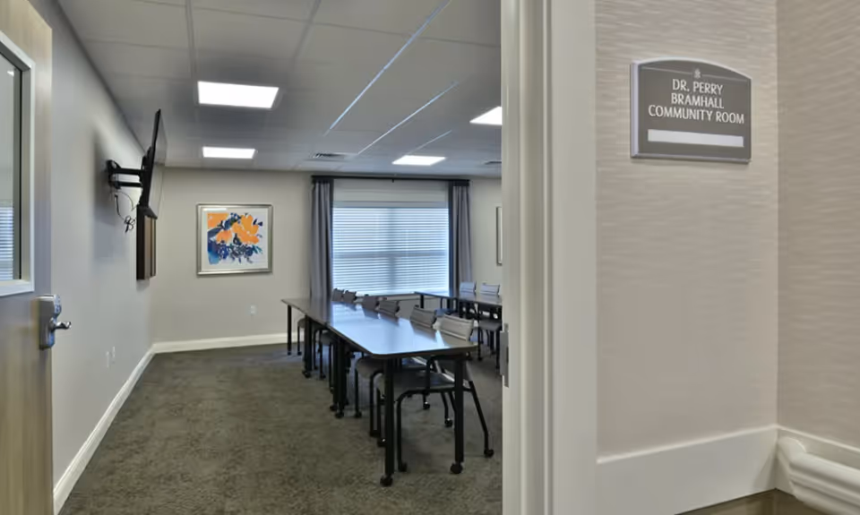 View into a community room with long tables and chairs, a wall-mounted TV, framed artwork, and a window with blinds.