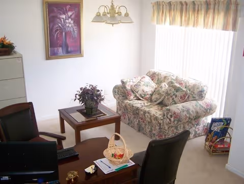 A small living room area with a floral-patterned loveseat, a wooden coffee table with a potted plant, and two black office chairs around a wooden desk. There is a framed floral painting on the wall, a window with vertical blinds and a valance, and a filing cabinet in the corner.