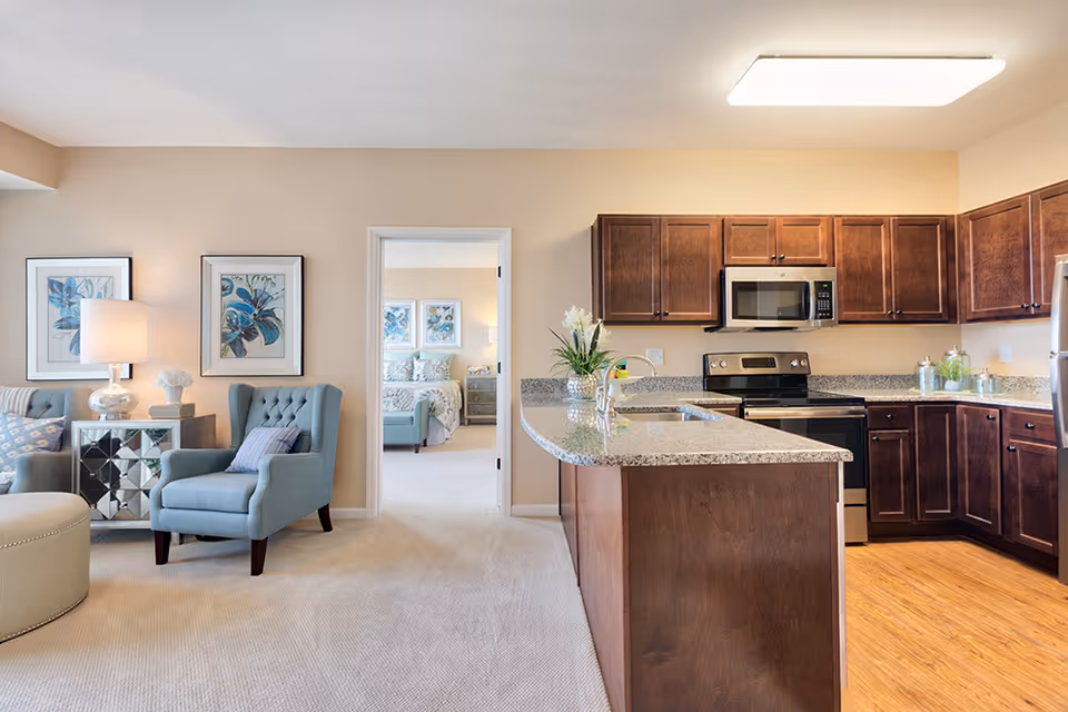 Interior view of a senior living facility apartment showing a living area with two blue armchairs, a side table with a lamp and decorative items, and framed artwork on the wall. Adjacent to the living area is a kitchen with dark wood cabinets, granite countertops, a stainless steel stove, microwave, and refrigerator. A doorway leads to a bedroom with a bed and matching artwork on the walls.