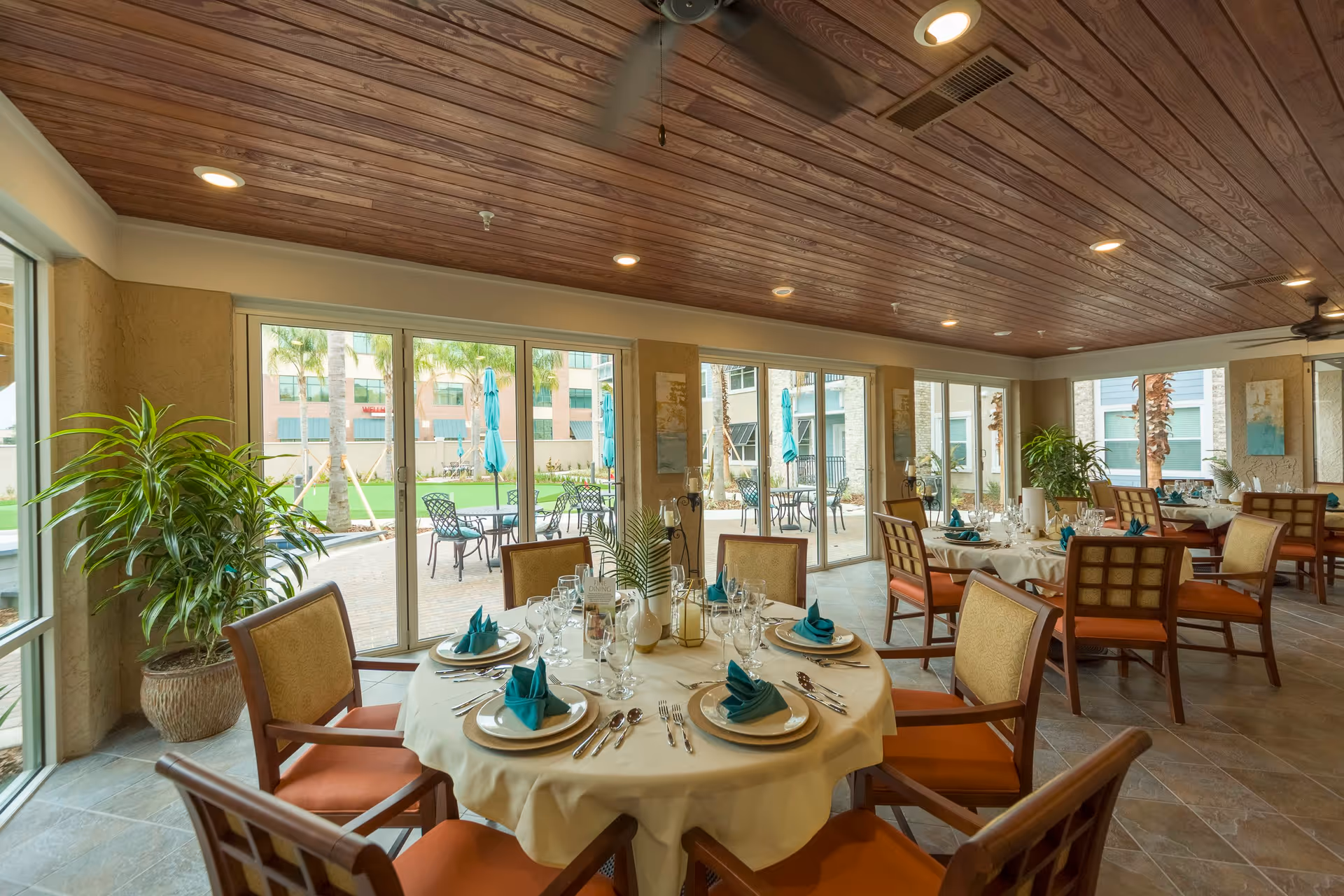 A bright dining room with round tables set with white tablecloths, plates, silverware, glasses, and teal napkins. The room has large windows and sliding glass doors that open to an outdoor patio with tables and umbrellas. The ceiling is wooden with recessed lighting and ceiling fans. There are potted plants inside near the windows.