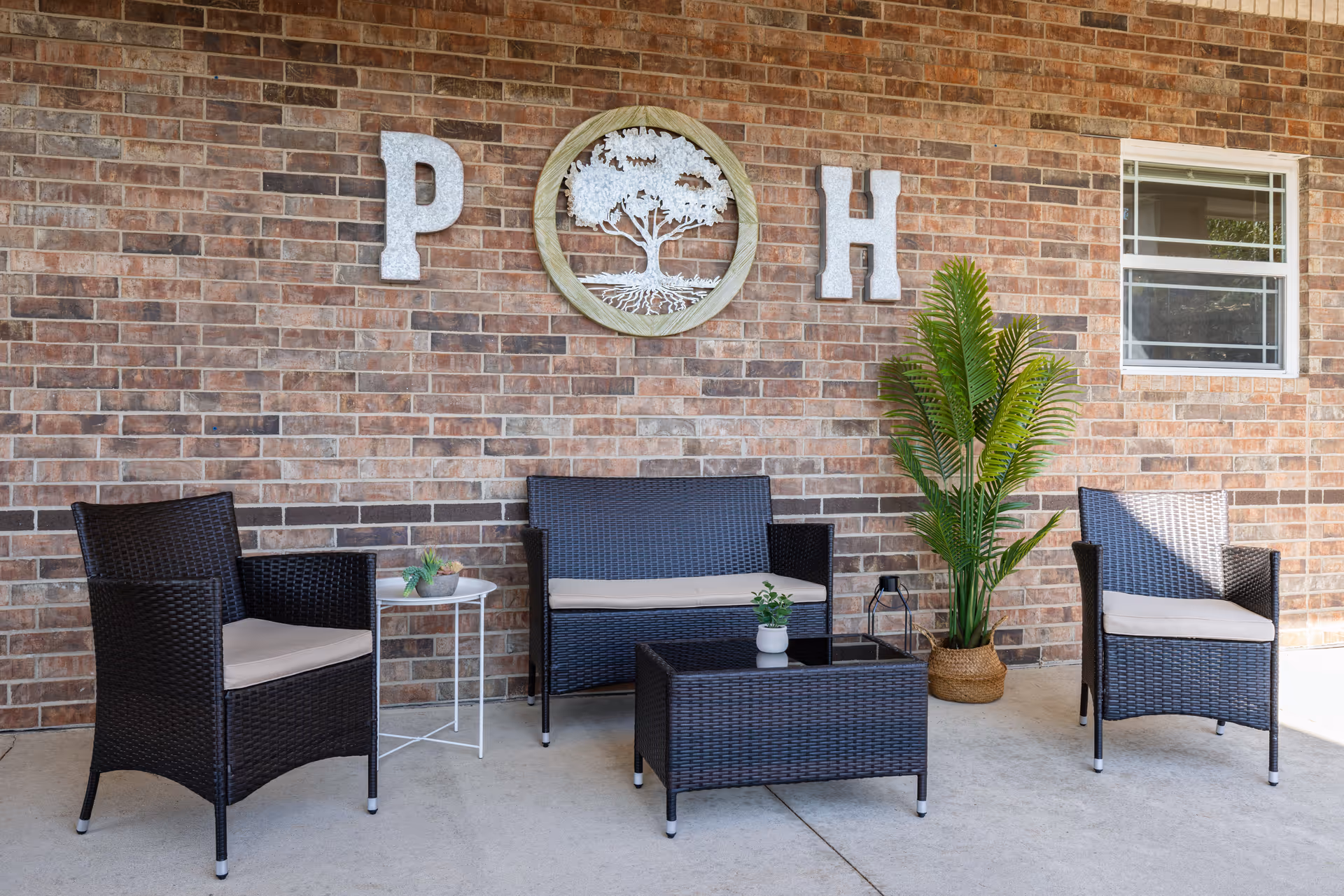 Covered brick patio with wicker seating, a coffee table, potted plant, and decorative wall letters 'P' and 'H' around a tree emblem.