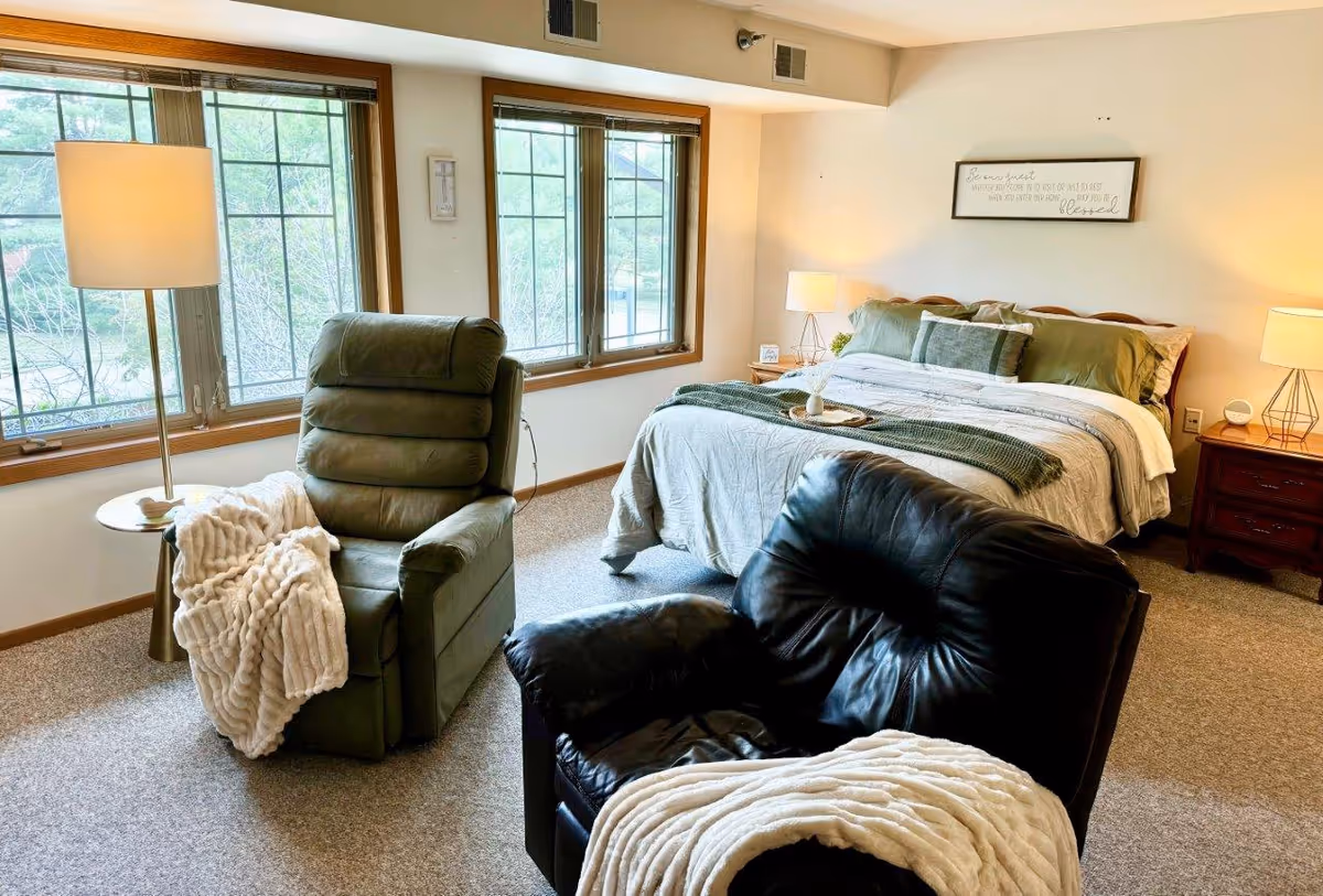 Well-lit bedroom with a made bed, two recliner chairs, large windows, and bedside tables with lamps.