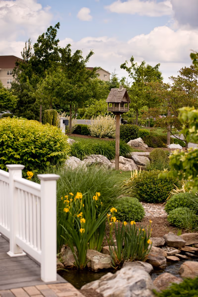 Well-maintained garden with a wooden birdhouse on a post, yellow flowers, rocks, and a white railing by a small pond.