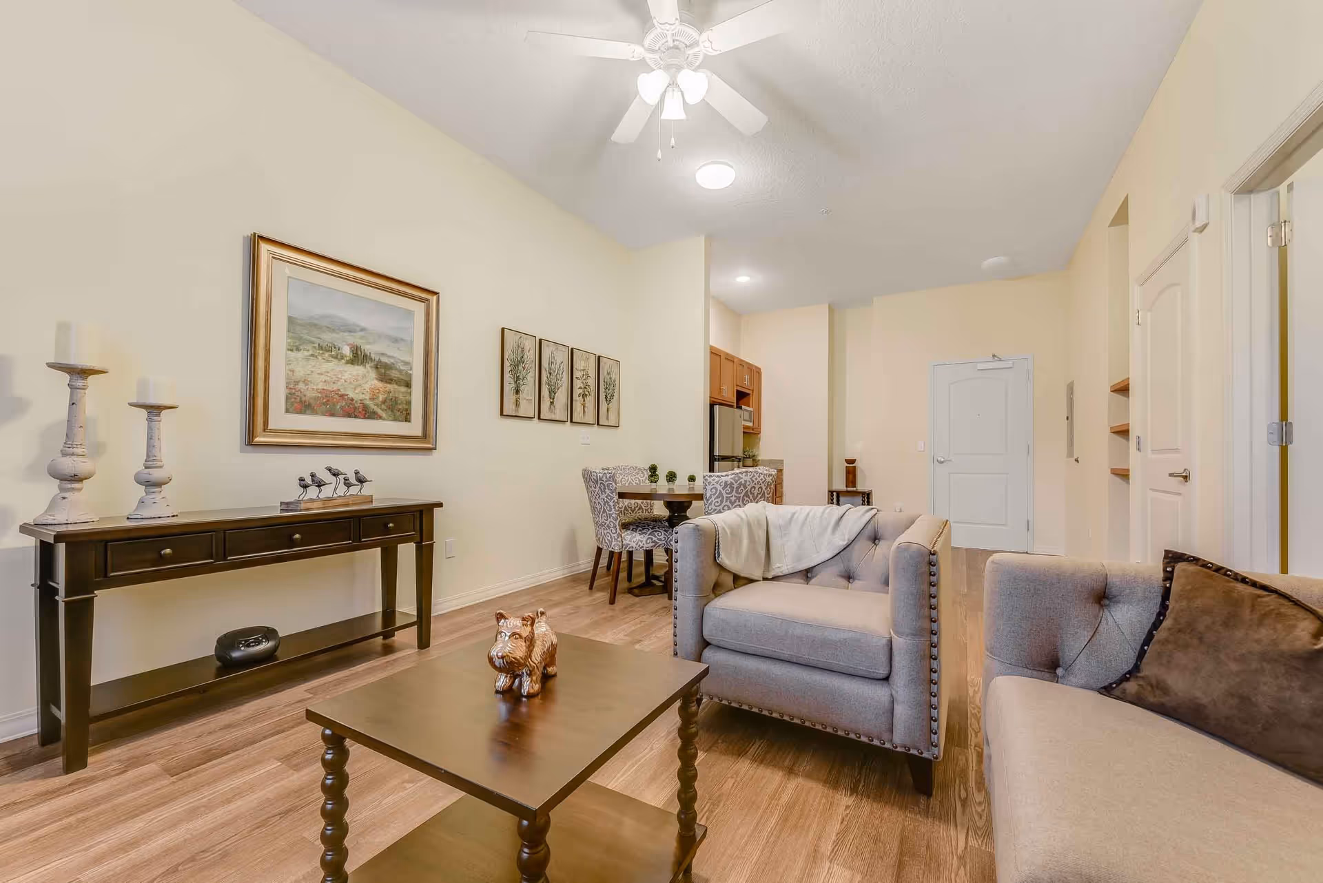 A cozy senior living room with beige walls and wooden flooring. The room features a gray armchair and a matching sofa with a brown pillow. A wooden coffee table with a small dog figurine is in the center. On the left wall, there is a dark wooden console table with two white candles and a bird decoration. Above the console table are framed paintings. In the background, there is a small dining area with a round table and four patterned chairs, and a kitchenette with wooden cabinets and stainless steel appliances. A ceiling fan with lights is mounted on the ceiling.
