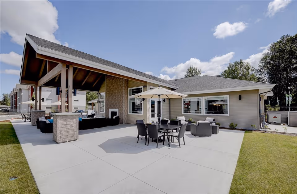Outdoor patio area at LARC at Kent featuring a covered seating area with cushioned chairs and a fireplace, an uncovered dining table with an umbrella, and additional seating arrangements on a concrete patio surrounded by grass and trees under a partly cloudy sky.