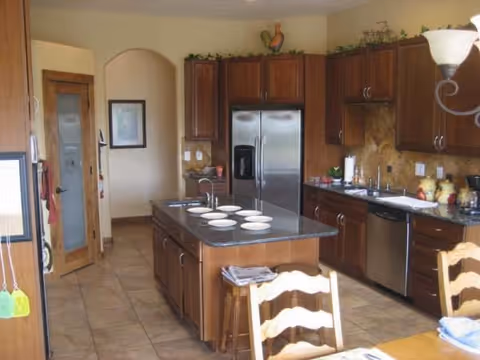 A spacious kitchen with a central island, wooden cabinets, stainless steel refrigerator and appliances, and a dining table with chairs.