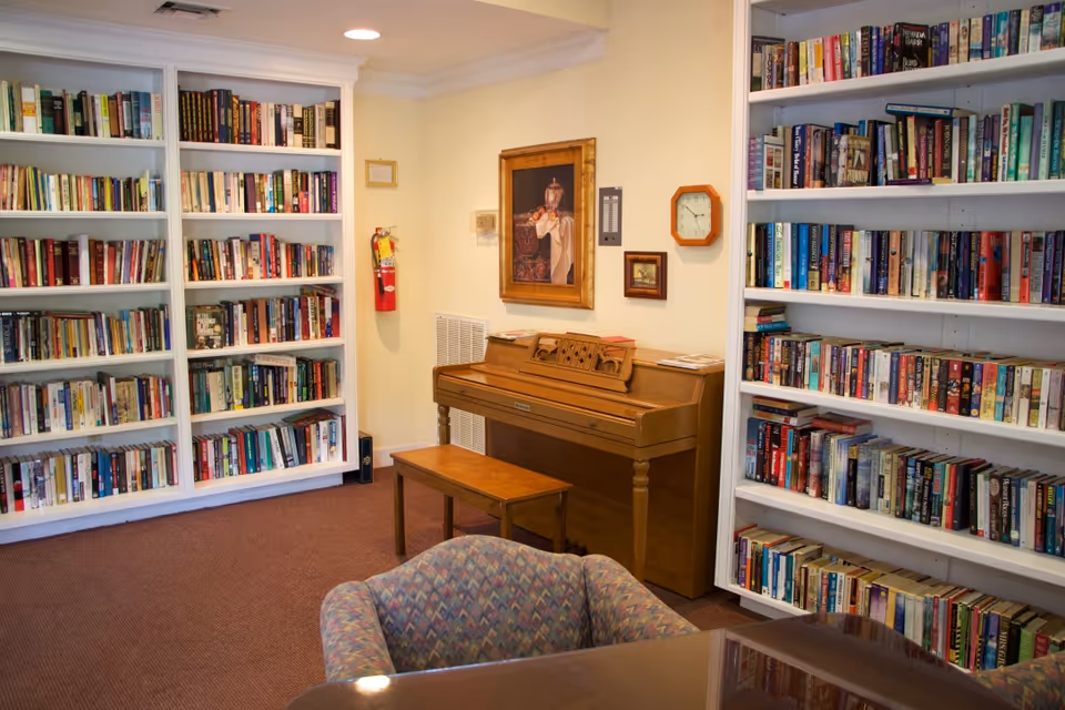 A cozy reading room with wall-to-wall bookshelves, a wooden piano and bench, and upholstered chairs.