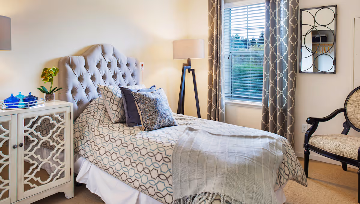 Well-decorated bedroom with a tufted headboard, patterned bedding, bedside cabinet, floor lamp, window with patterned curtains, and an armchair.