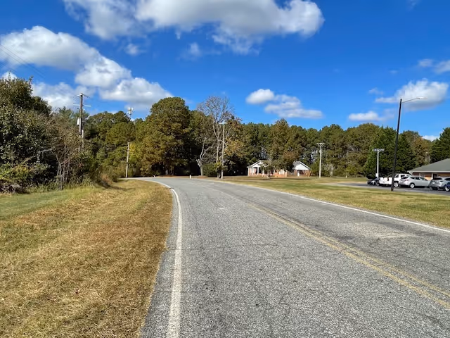 Two-lane paved road curving through a grassy rural area with trees and a few buildings under a blue sky with scattered clouds.