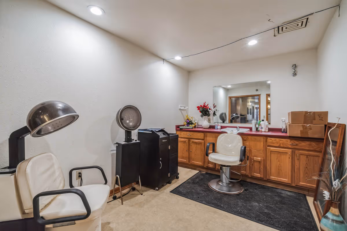 Interior view of a small salon area with two white salon chairs, one under a hair dryer and another in front of a large mirror on a wooden counter with cabinets. Various hair care products and boxes are on the counter, and the room has white walls and recessed ceiling lights.