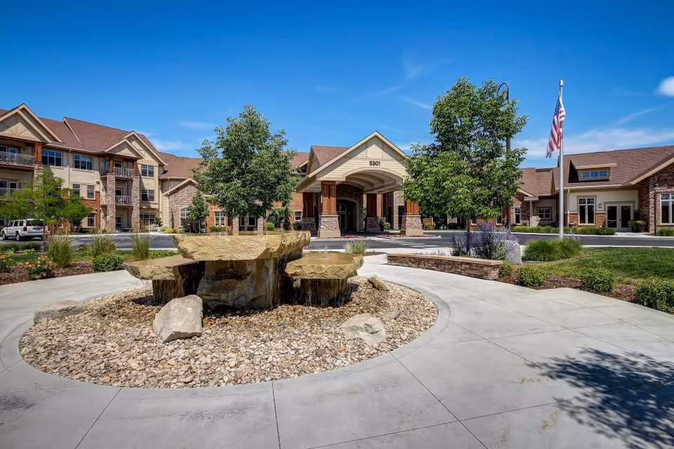 Stone tiered fountain in a circular plaza in front of the covered entrance to a multi-story senior living building with an American flag.