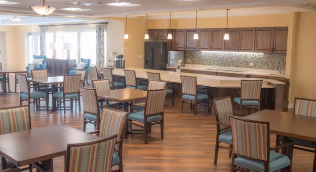 A spacious dining area with multiple wooden tables and chairs with striped upholstery. In the background, there is a kitchen area with a long counter, pendant lights, dark wood cabinets, a refrigerator, and a mosaic tile backsplash. Large windows with curtains allow natural light to fill the room.