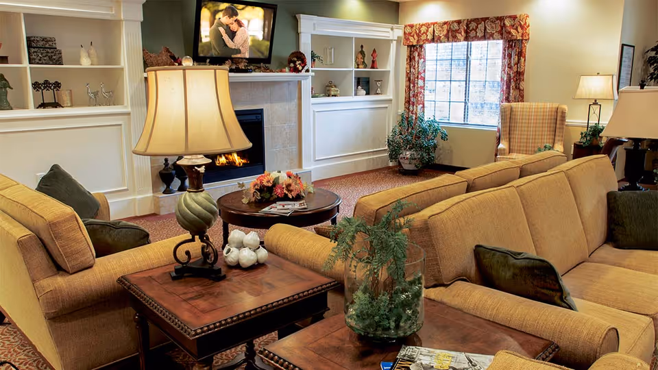 A cozy living room with beige sofas and green cushions arranged around a wooden coffee table and side tables. A lit fireplace is set in a white mantel with built-in shelves on either side displaying decorative items. A flat-screen TV above the fireplace shows a couple embracing. A window with floral curtains lets in natural light, and there are several lamps and plants adding warmth to the room.